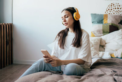 Pregnant attractive woman sitting on the bed in headphones looks at social networks on a mobile.