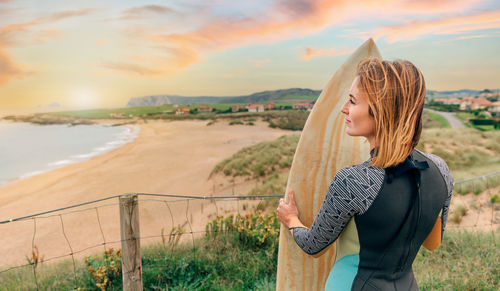 Surfer woman with wetsuit and surfboard looking at the beach