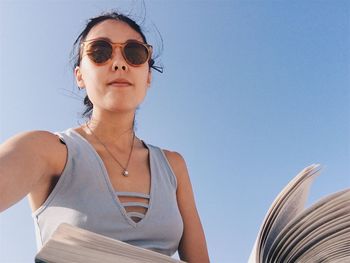 Low angle portrait of young woman holding book against clear blue sky