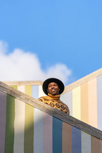 From below of african american male in trendy cap and warm knitted sweater standing on steps of staircase against striped colorful wall and looking away