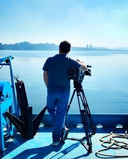 Rear view of man with camera on tripod standing by sea against sky