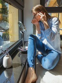 Young beautiful girl applies makeup on her face next to the window. reflection in the mirror