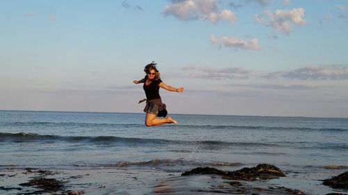 Full length of mature woman jumping with arms outstretched on shore at beach against sky