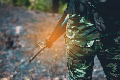 Midsection of person in camouflage clothing holding rifle while standing outdoors
