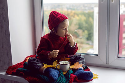 Boy a child in a red sweater and a knitted hat sits at the window on the windowsill