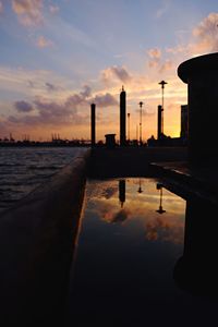 Silhouette factory by sea against sky during sunset