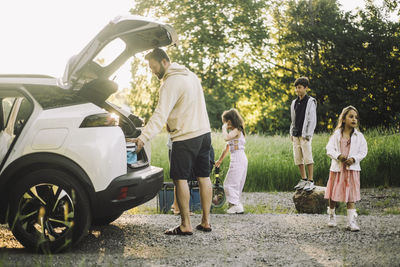 Father unloading stuffs from electric car trunk on dirt road