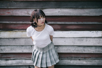 Young woman looking away while standing against wooden wall