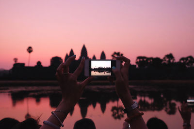 Close-up of cropped hand photographing angkor wat by lake at sunset