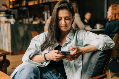 Young brunette woman sitting in coffee shop uses mobile phone for online messages.