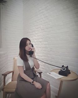 Young woman using telephone while sitting on chair against wall