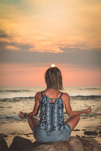Woman doing yoga on rock at beach against sky during sunset