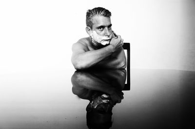 Portrait of man shaving while sitting with reflection on table against white background