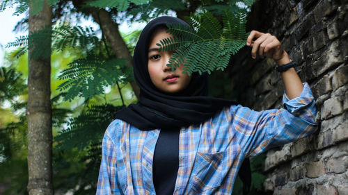 Portrait of woman holding leaves over face while standing by wall against tree