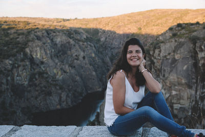 Portrait of smiling young woman sitting on rock formation against sky