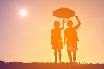 Full length of silhouette friends holding umbrella while standing against sky during sunset