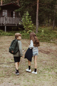 Full length of brother and sister with bag standing against house in forest