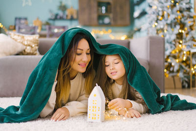Mom and daughter are lying under a blanket and looking at a glowing toy house