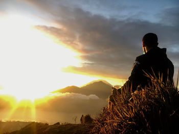 Low angle view of young man sitting on field against cloudy sky during sunset