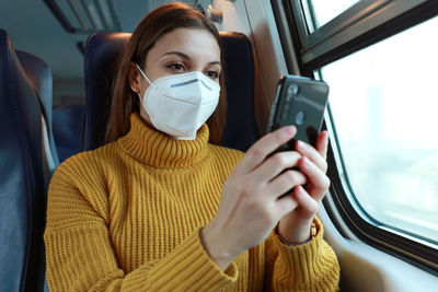 Young woman in mask using mobile phone while sitting at bus