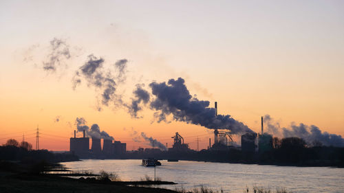 Smoke emitting from factory by sea against sky during sunset