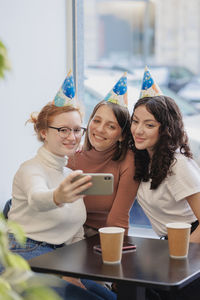 Smiling woman wearing birthday cap taking selfie with friends at cafe