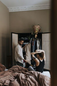 Man showing dress to non-binary person tying hair while sitting on bed at home