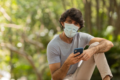 Young man wearing a mask and a smartphone at day time at a green park . mobile phone, technology