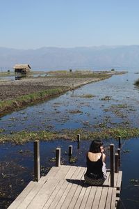 Rear view of woman sitting by lake against sky