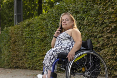 Smiling young woman with short stature on wheelchair talks on smartphone in green park at summer day