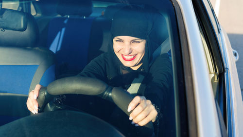 Smiling woman wearing hijab screaming while driving car seen through windshield