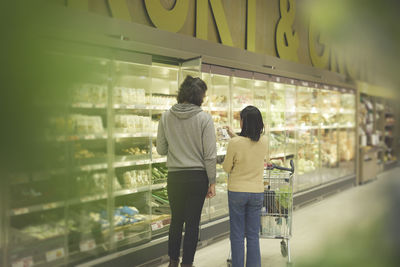 Rear vie of couple standing in front of fridges with vegetables while doing shopping in supermarket