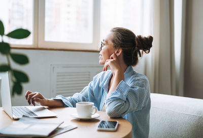 Young smiling woman in blue shirt using laptop drinking tea in room