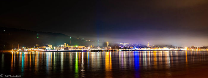 Illuminated city by sea against sky at night