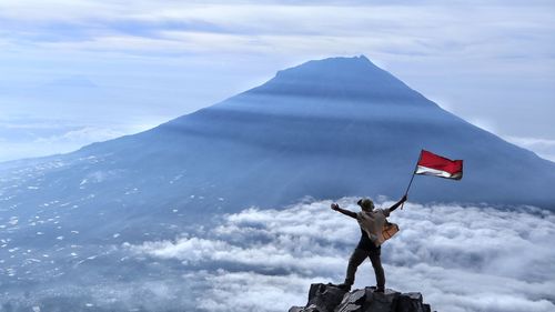 Rear view of man holding flag while standing on rock against sky