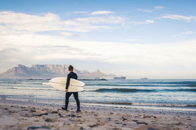 Rear view of a surfer standing at beach against sky with table mountain in south africa 