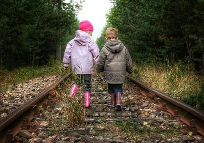 Rear view of siblings wearing hooded jacket walking on railroad track amidst trees