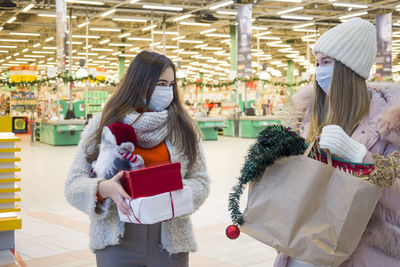 Smiling young women wearing mask holding christmas gifts standing at supermarket