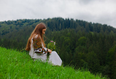 Girl in embroidery with a bouquet of wild flowers sits on green grass in the carpathian mountains