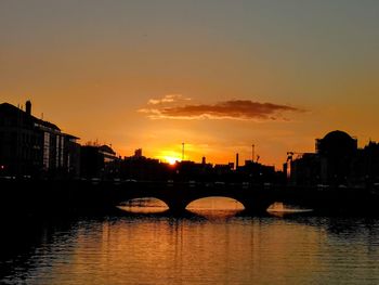 Bridge over river by silhouette buildings against sky during sunset