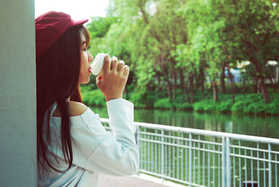 Side view of young woman having drink by railing against trees