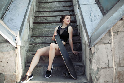 Full length of beautiful woman holding skateboard while sitting on staircase
