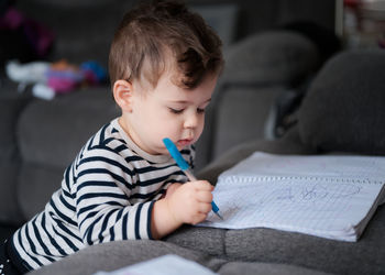 Young baby boy toddler writing in a notebook on the couch at home