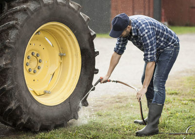 Full length of man washing tractor wheel with hose in farm