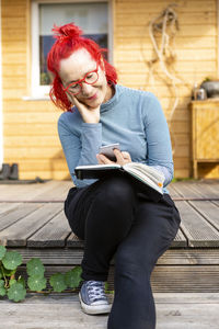Portrait of smiling senior woman with red dyed hair sitting on terrace in front of her house using cell phone