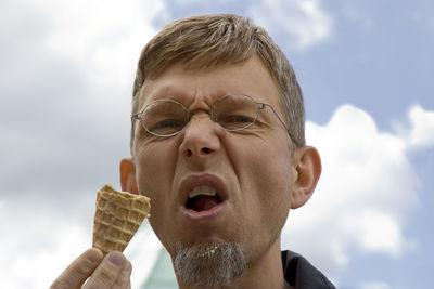 Low angle portrait of mature man making face while having ice cream cone against sky