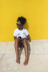 Full length of young woman sitting with mobile phone against yellow wall