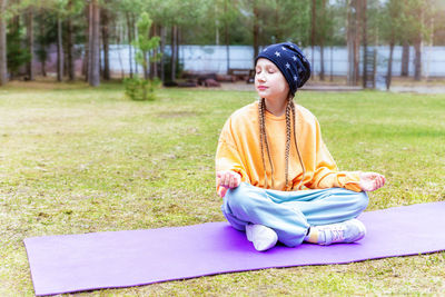 Beautiful caucasian teenage girl sitting on a sports mat outdoors, meditating with her eyes closed. 