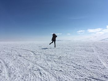 Full length of young woman jumping on snow covered field against blue sky