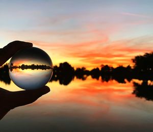 Cropped hand holding crystal ball over lake during sunset